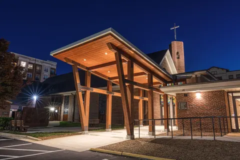 Accessible ramp and walkway leading under the illuminated timber canopy toward the brick bell tower at Ascension Church in Charlotte, NC, at night, by Ossa Studio.