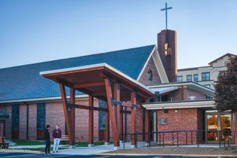Brick bell tower and cross rising behind the new timber canopy and accessible walkway at Ascension Church in Charlotte, NC, a church entry project by Ossa Studio.