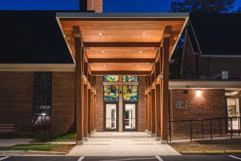 Head-on night view beneath the Ascension Church canopy in Charlotte, NC, where downlights in the wood ceiling lead to stained-glass entry doors, an Ossa Studio design.