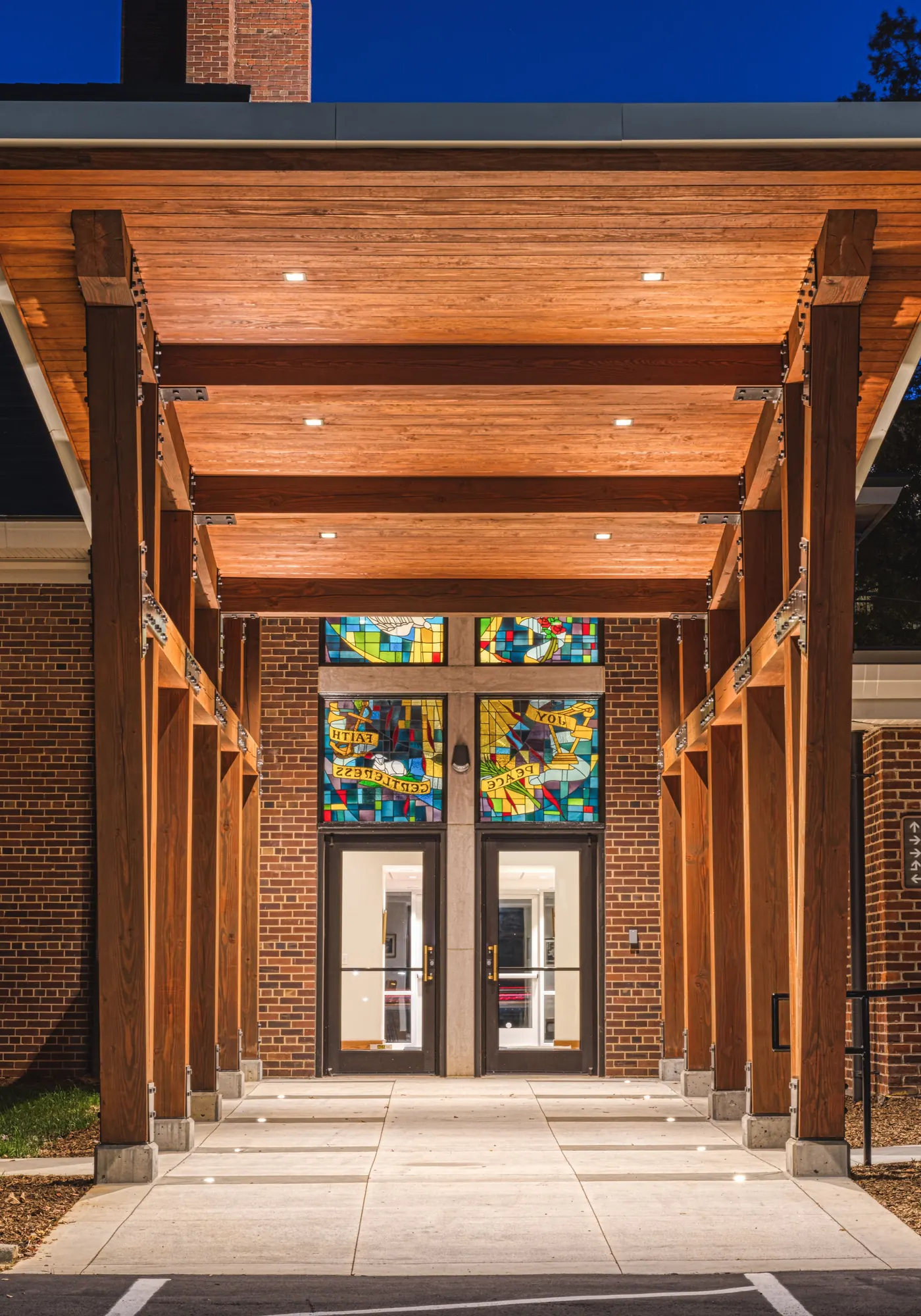 Vertical night view down the lit timber entry canopy at Ascension Church in Charlotte, NC, with rows of wood columns and a warm ceiling leading to stained-glass doors, a church entry designed by Ossa Studio.