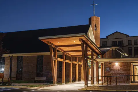 Timber entry canopy lit at night in front of the brick sanctuary and bell tower at Ascension Church in Charlotte, NC, designed by Ossa Studio.