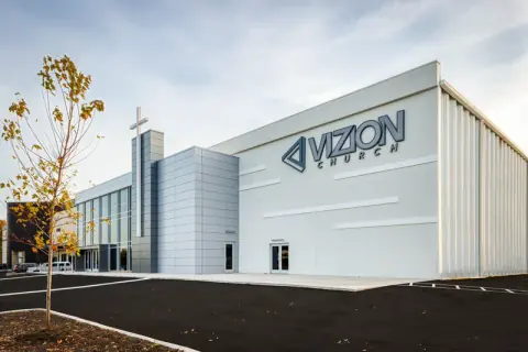 Side elevation of Vizion Church in Charlotte, NC, with large wall signage, a metal-panel cross tower and the glass entry wing on the renovated commercial building, an adaptive reuse by Ossa Studio.