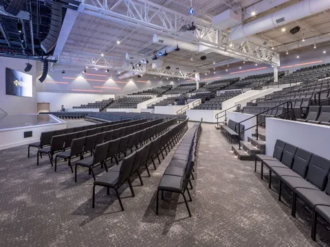 Rows of floor seating and raised seating tiers under the exposed roof trusses and lighting rigs of the Vizion Church auditorium in Charlotte, NC, by Ossa Studio.