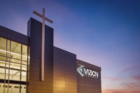 Close view of the metal-panel cross tower beside the lit glass lobby and wall signage at Vizion Church in Charlotte, NC, at dusk, by Ossa Studio.