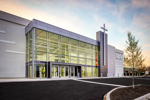 Angled dusk view of the lit two-story glass lobby and cross tower at the Vizion Church entrance in Charlotte, NC, part of the church conversion by Ossa Studio.