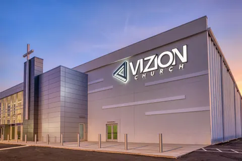 Corner view at dusk of the illuminated Vizion Church sign, cross tower and metal-panel walls on the converted building in Charlotte, NC, designed by Ossa Studio.